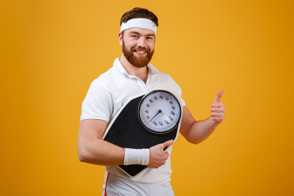 رجل مبتسم يحمل ميزانًا تعبيرية عن إيجاد طريقة لخسارة الوزن التدريجية مناسبة لنمط حياتك